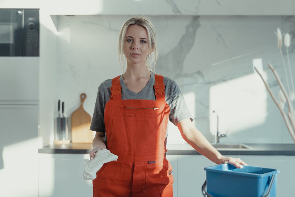 A professional cleaner from Cleaners TW1 stands in a modern kitchen holding a white cleaning cloth in one hand and resting the other hand on a blue cleaning bucket. The countertop is made of sleek, white material with a stainless steel sink and a chrome faucet. Behind the cleaner, there is a white marble backsplash with grey veining, and a wooden chopping board is propped against the wall. The room is brightly lit with natural light, highlighting the spotless, shiny surfaces, including a wooden cutting board and stainless steel appliances, indicating a thorough surface cleaning process. The cleaner is dressed in a grey t-shirt and bright orange apron, emphasizing her role in domestic cleaning and sanitization services offered by Cleaners TW1, visible through the natural and professional presentation aligned with affordable end of tenancy cleaning in TW1.