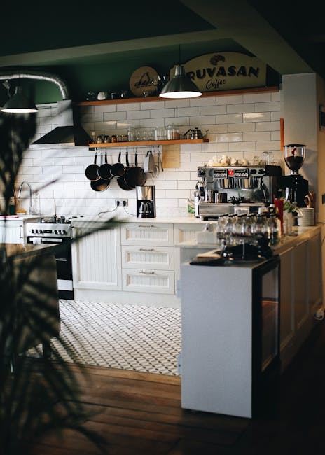A modern kitchen featuring a white subway tile backsplash and white cabinetry with silver handles. The countertop is clutter-free and appears clean, with a professional espresso machine and glass jars neatly arranged on it. Above the countertop, there is a wooden shelf holding cups and small containers, with black pots hanging from hooks beneath. Stainless steel appliances, including a stove with a range hood, are visible to the left. The floor has a patterned rug, and the room is illuminated by professional lighting, creating a bright and hygienic atmosphere suitable for deep cleaning and sanitisation services, as offered by Cleaners TW1 near Twickenham Station, TW1.