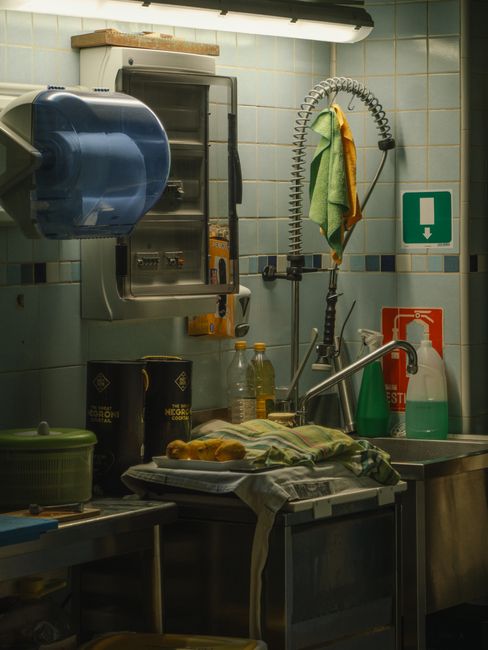 A commercial kitchen sink area featuring stainless steel surfaces and a tiled wall in pale blue and white. The deep sink has a flexible, coiled spray hose with a yellow cloth hanging from it. To the left, there are several bottles, including a clear plastic bottle and two black containers labeled 'The Mercurial Cook'. On the stainless steel countertop, a white dishcloth is draped over a pile of dishes, and a green plastic container with a lid is visible. Above, a wall-mounted paper towel dispenser and an emergency shower sign are present, alongside wall-mounted safety and fire equipment indicators. The scene is illuminated by overhead fluorescent lighting, emphasizing the cleanliness of the surfaces, which appear well-maintained and ready for sanitisation and surface cleaning tasks in line with deep cleaning standards for kitchens near Twickenham Station, TW1. Cleaners TW1 ensure these areas meet hygiene and cleanliness requirements regularly, adhering to professional cleaning practices.