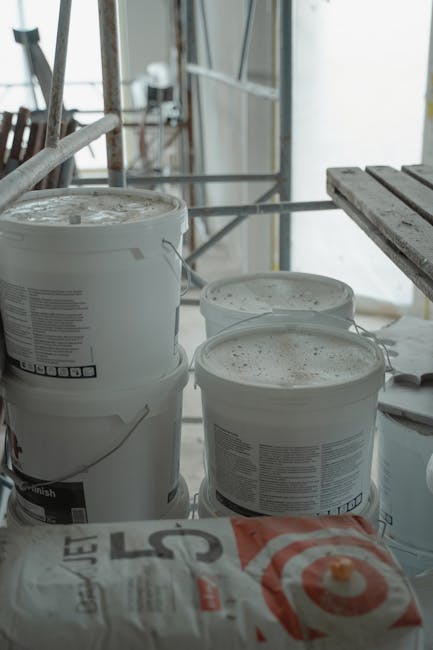 An indoor construction site during a building renovation, featuring three white plastic containers with lids placed on the floor, surrounded by scaffolding and construction materials. The surfaces of 