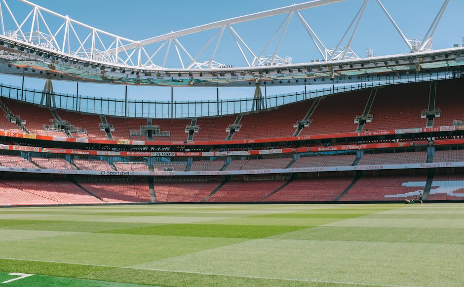 An empty stadium featuring a well-maintained green grass pitch with alternating light and dark stripes, surrounded by red tiered seating sections. The seating is arranged in curved rows with black railings and various advertisements displayed on the perimeter. Above, a white modern roof structure with a geometric design extends across the top of the stadium, supported by white beams, against a clear blue sky. The overall scene is bright, clean, and shows no signs of dust or debris, exemplifying professional cleaning standards suited for large-scale venues like Twickenham Stadium, as promoted by Cleaners TW1 for same-day emergency cleaning services.