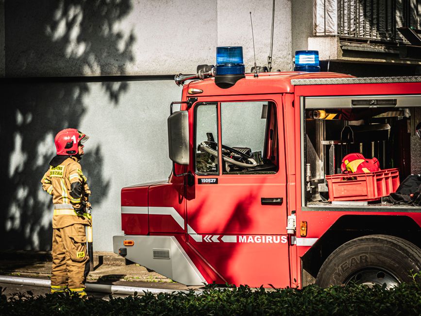 A firefighter dressed in yellow protective gear and a red helmet stands beside a red fire engine with blue emergency lights on the roof, parked outdoors near a concrete wall, during daytime. The scene is illuminated by natural sunlight, casting shadows on the wall and ground. The fire engine has visible compartments and equipment storage on its side, with a clear view of its large black tire and reflective striping. The setting suggests a readiness for emergency response, with the firefighter preparing or inspecting the vehicle, which is part of routine safety and emergency services organized by Cleaners TW1 in relation to the Twickenham Stadium emergency cleaning services page.